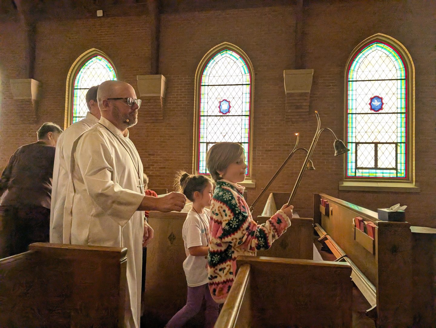 Photo of four people walking down the center aisle of the church. The two figures in the front are children holding long candles on poles followed by two men in white robes.